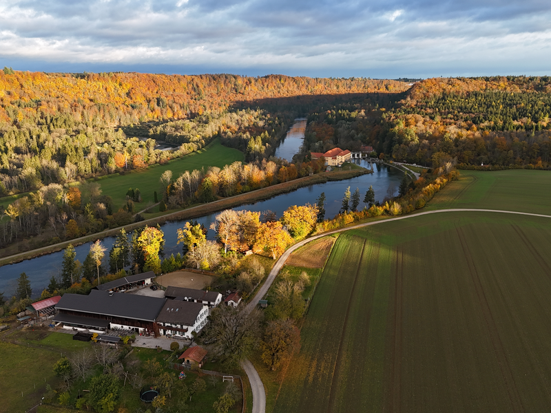 Eine herbstliche Landschaft mit einem Fluss, der durch bewaldete Hügel fließt, während im Vordergrund ein großes Gebäude mit umgebenden Feldern zu sehen ist.