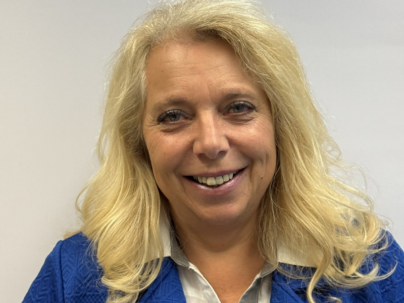 Eine blonde Frau mit langen Haaren lächelt in die Kamera, gekleidet in einem blauen Oberteil vor einem neutralen Hintergrund.
