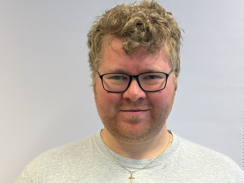 Ein Mann mit lockigem blondem Haar, Brille und einem Kettchen mit Kreuz lächelt in die Kamera.