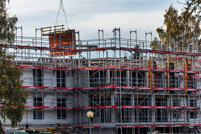Auf dem Bild ist ein teilweise gebautes Gebäude mit einem Gerüst zu sehen, an dem ein Kran ein Bauteil hebt.